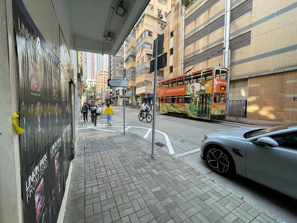 Ref#3607 shows a city street scene with pedestrians on the sidewalk, a cyclist passing by, and an orange double-decker tram. Tall buildings with rental signs line the street, and a silver car is parked in the foreground.