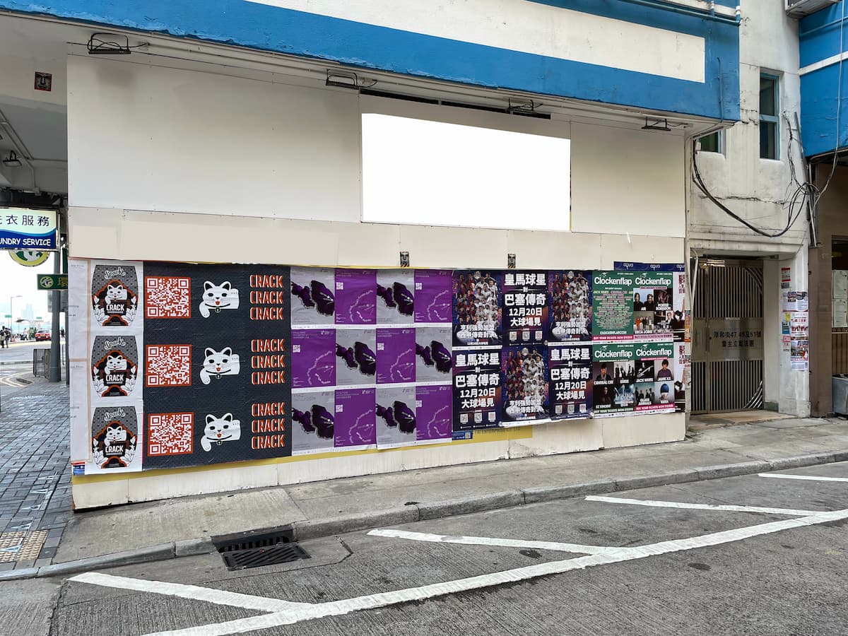 A white building wall with colorful posters in purple, black, red, and green stands by a gated entrance and city sidewalk, suggesting nearby rental options for office or shop space in Ref#36079s lively urban location.
