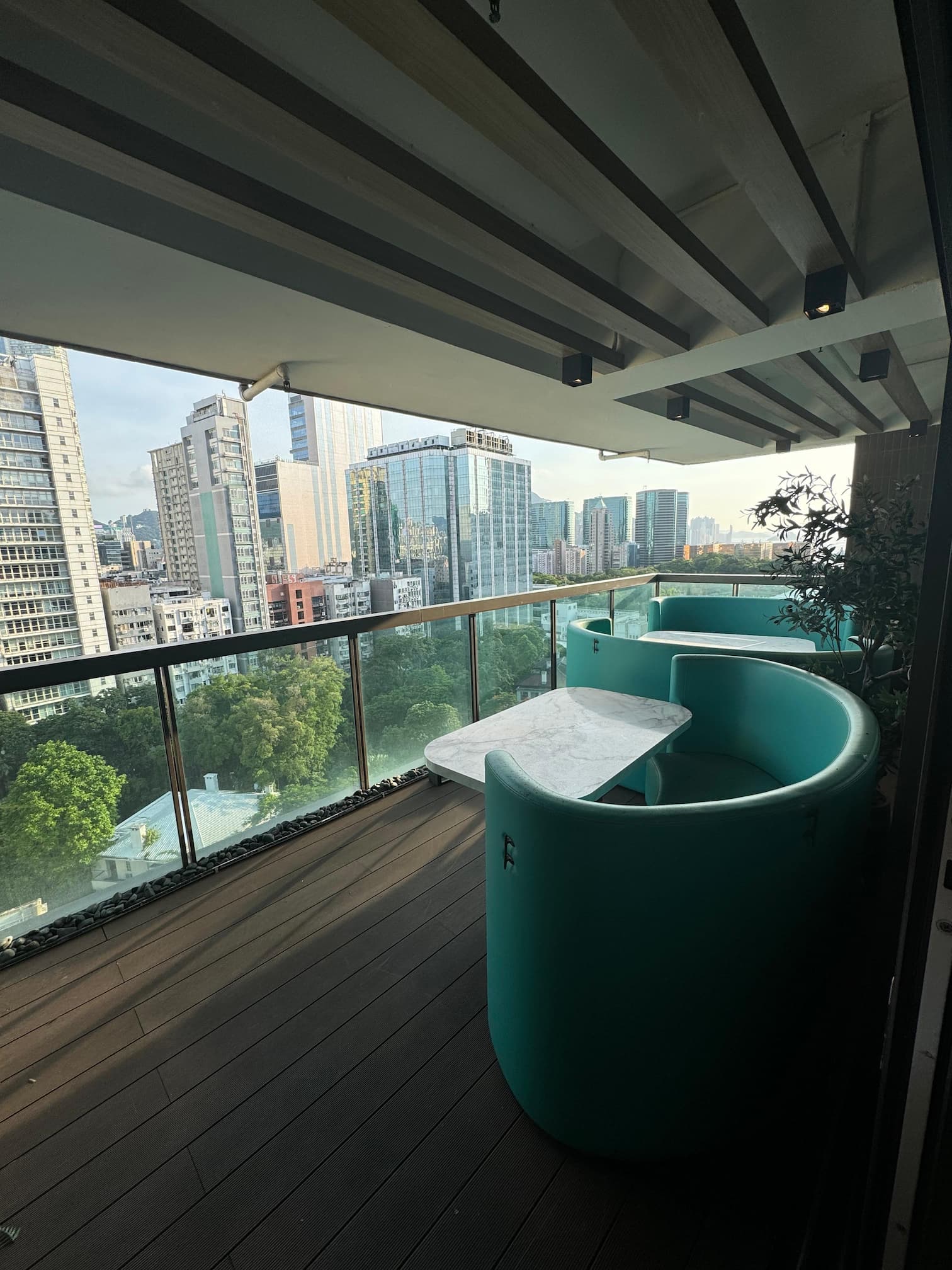Ref#3612: A modern balcony, perfect for office rental, showcases a curved turquoise sofa and white marble table with views of city skyscrapers and green trees beneath a partly cloudy sky.