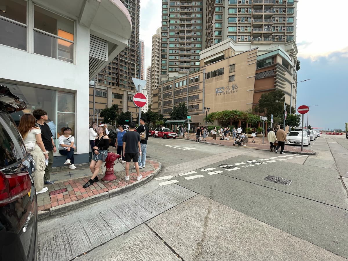A group of people gather on a city sidewalk near tall apartments and several parked cars. Red and white no entry signs are visible. This scene is featured in product Ref#3608.