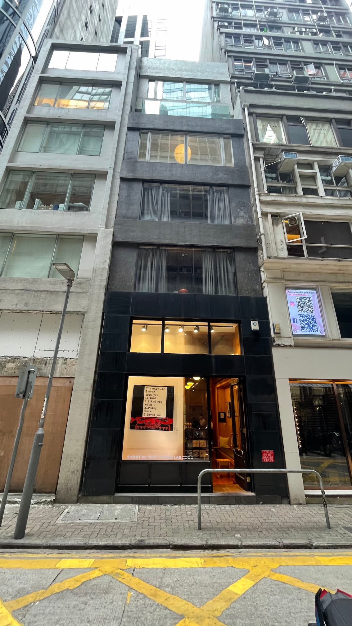 Ref#3600 is a narrow, multi-story building with a black facade and large windows on a city street. The brightly lit ground floor entrance displays signage, making it ideal for office or shop rentals amidst surrounding tall buildings.
