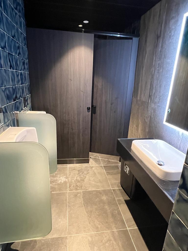 Ref#3602: A modern restroom ideal for rental restaurants or offices, with two urinals with frosted dividers, a closed stall with dark wood door, rectangular sink, backlit mirror, and tiled floor and walls for easy maintenance.