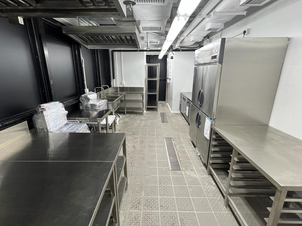 Ref#3602: Commercial kitchen for restaurant rental with stainless steel counters, shelves, appliances (including a large refrigerator), tiled floors, and organized space with trays and chairs stacked along one wall.