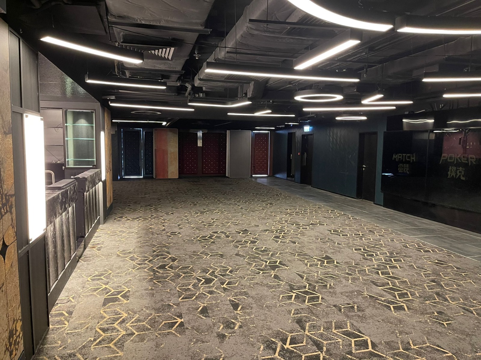 Ref#3602 is a spacious, modern room with patterned carpet, geometric ceiling lights, dark walls, and glass panels labeled MATCH and POKER1ideal for office rental. The well-lit area is empty and features doors at the far end.