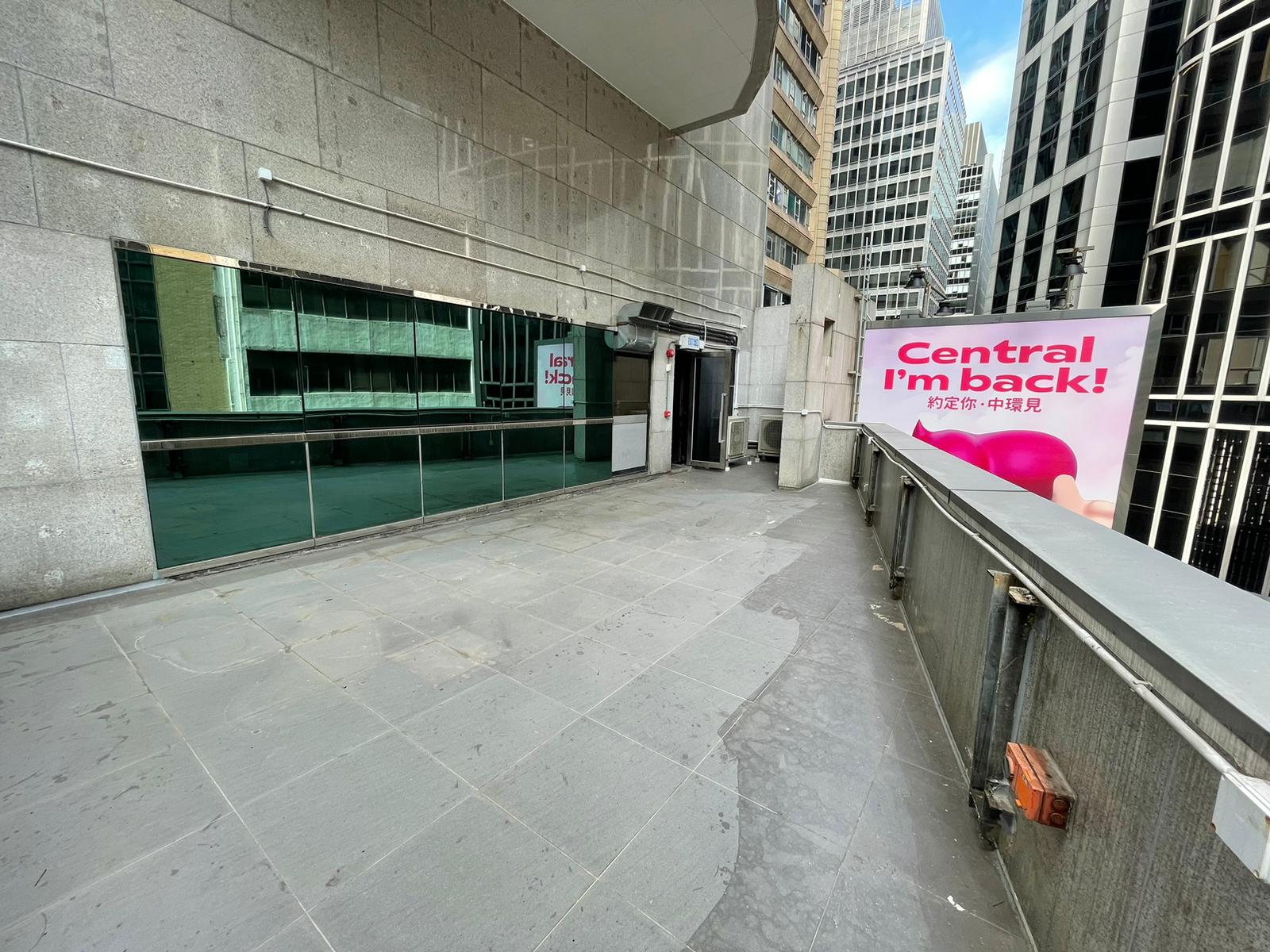 Ref#3602: City rooftop terrace with grey tiles, glass panels, and a tall pink "Central Im back!" sign in English and Chinese. Ideal for shop rental, surrounded by skyscrapers beneath a clear sky.
