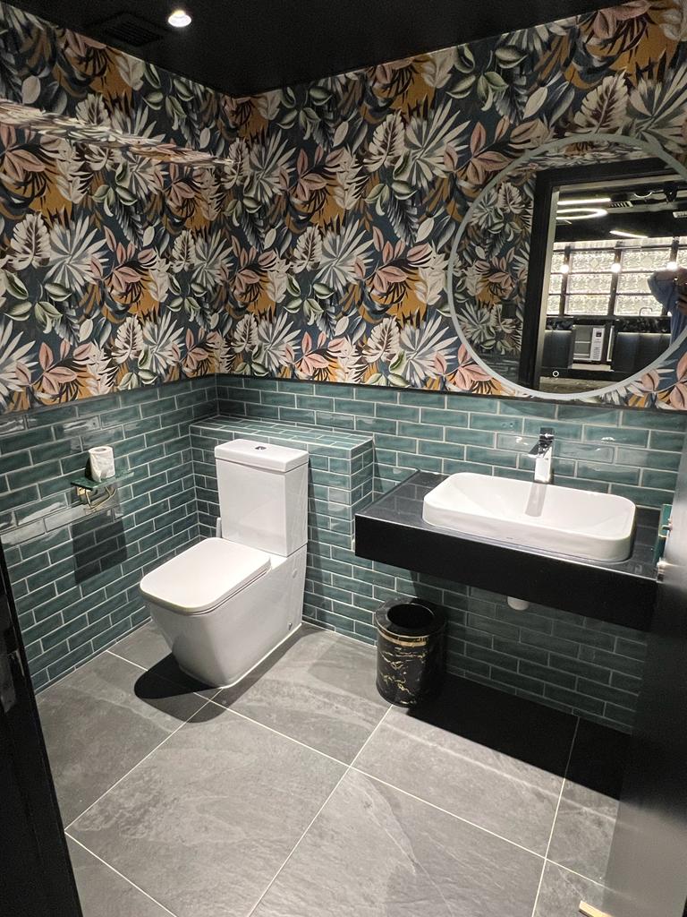 Ref#3602: A stylish shop rental bathroom with green subway tile walls, floral wallpaper, a white toilet, modern square sink on a black countertop, round mirror, and large gray floor tiles.