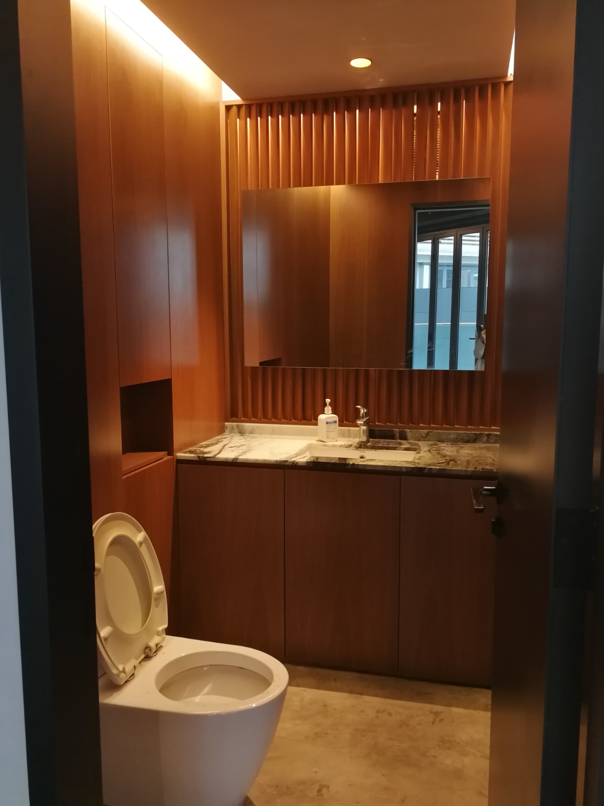 The modern bathroom in office rental Ref#3597 features wooden panel walls, an open white toilet, marble countertop with sink, soap dispenser, and a large mirror reflecting a window, all illuminated by warm lighting.