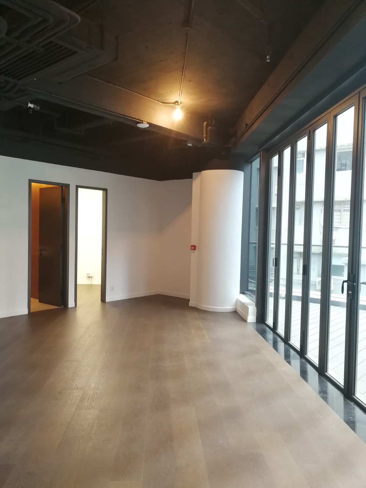 Ref#3597: Modern, empty office rental featuring dark wood floors, black ceiling, and floor-to-ceiling windows. Two open doorways on the left reveal a bright interior and a small white room.