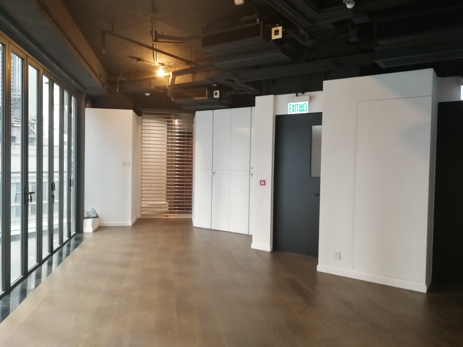 Ref#3597: Modern empty office with wooden floors, white walls, black ceiling, tall left-side windows013ideal for office or shop rental013features built-in cabinets and an illuminated exit sign above a dark doorway.