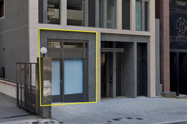 Ref#3248 is a small, modern shop rental with large glass double doors framed in yellow at the base of a tall building. The entrance sits next to a metal gate and a grey wall-mounted lamp.