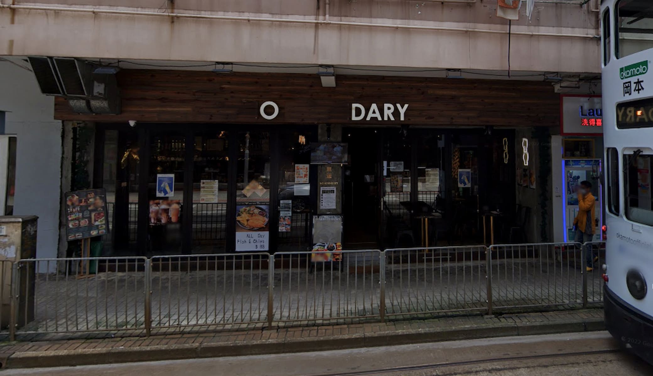 Ref#3240 is a dark storefront with large glass windows, a wooden facade, and partial "O DARY" signage
dical for a shop or restaurant. Posters and menus are displayed, and a metal fence lines the sidewalk in front.