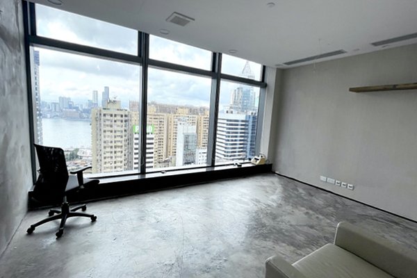 Ref#3281: A mostly empty office rental featuring a gray floor, black office chair, and couch facing floor-to-ceiling windows with views of a cityscape of tall buildings and a river beneath a cloudy sky.