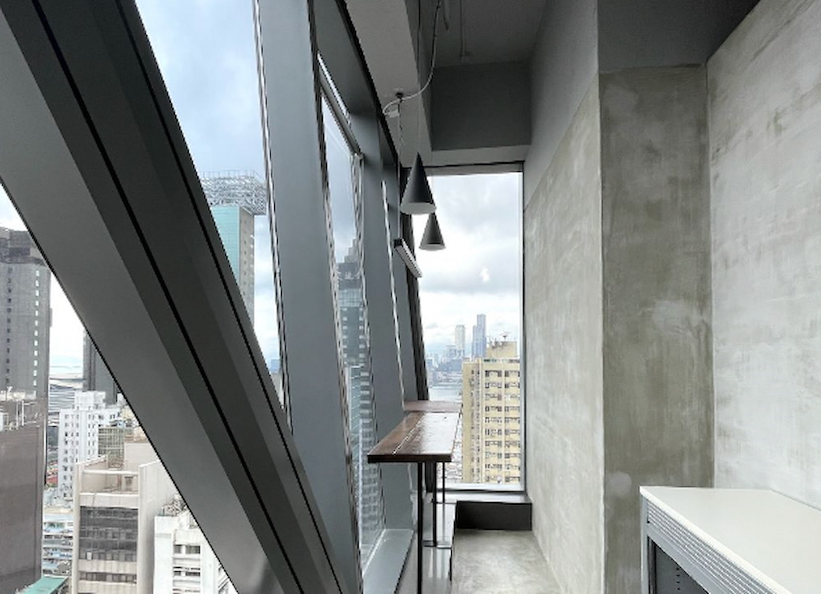 Ref#3281 is a modern high-rise office rental with slanted floor-to-ceiling windows, a narrow wooden desk, two pendant lights, and views of city buildings under a cloudy sky.