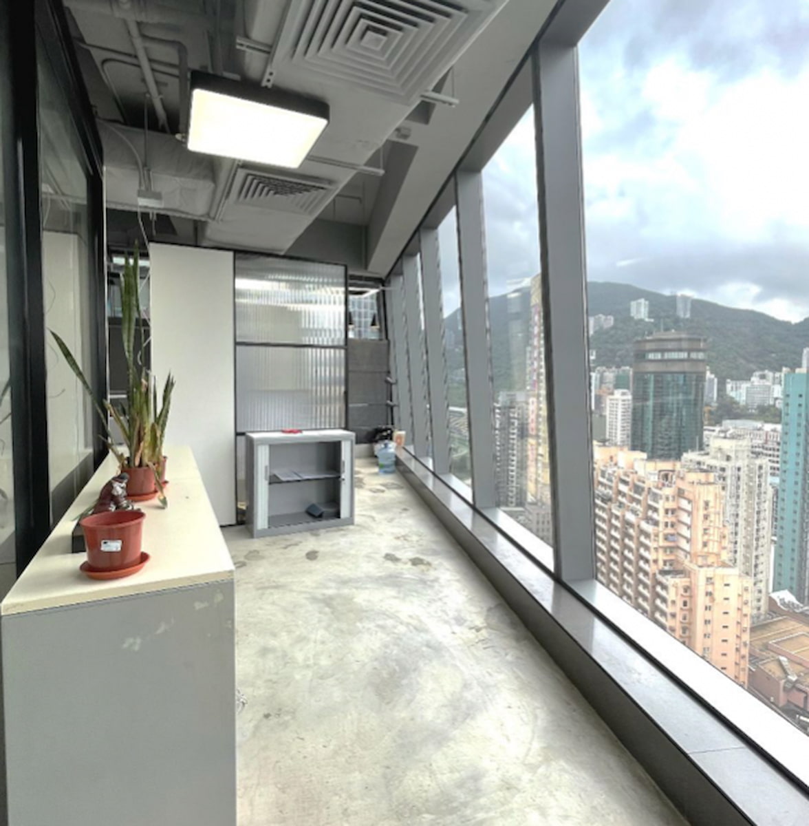 Ref#3281: Long corridor with floor-to-ceiling windows offering city and mountain views, ideal for office rental. Features potted plants and a small cabinet for a welcoming atmosphere.