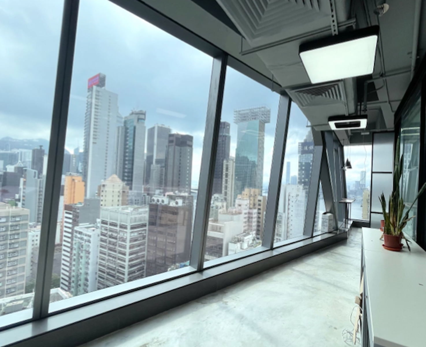 Ref#3281: Modern office rental interior with large floor-to-ceiling windows providing a panoramic city skyline view, tall buildings under a cloudy sky, and an indoor plant adding freshness on the counter by the window.