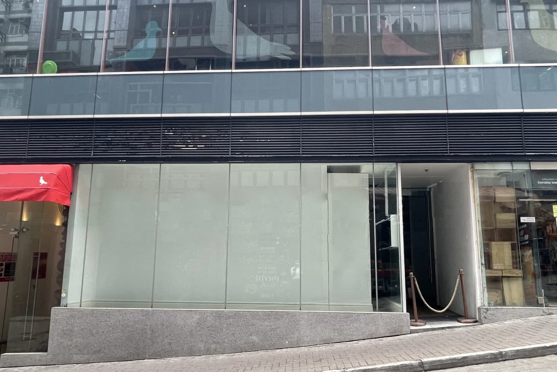 Ref#3355: A glass-fronted building with a partially frosted window and open glass door, ideal for office rental, features a rope barrier at the entrance, a red awning to the left, and sits on a sloped street.