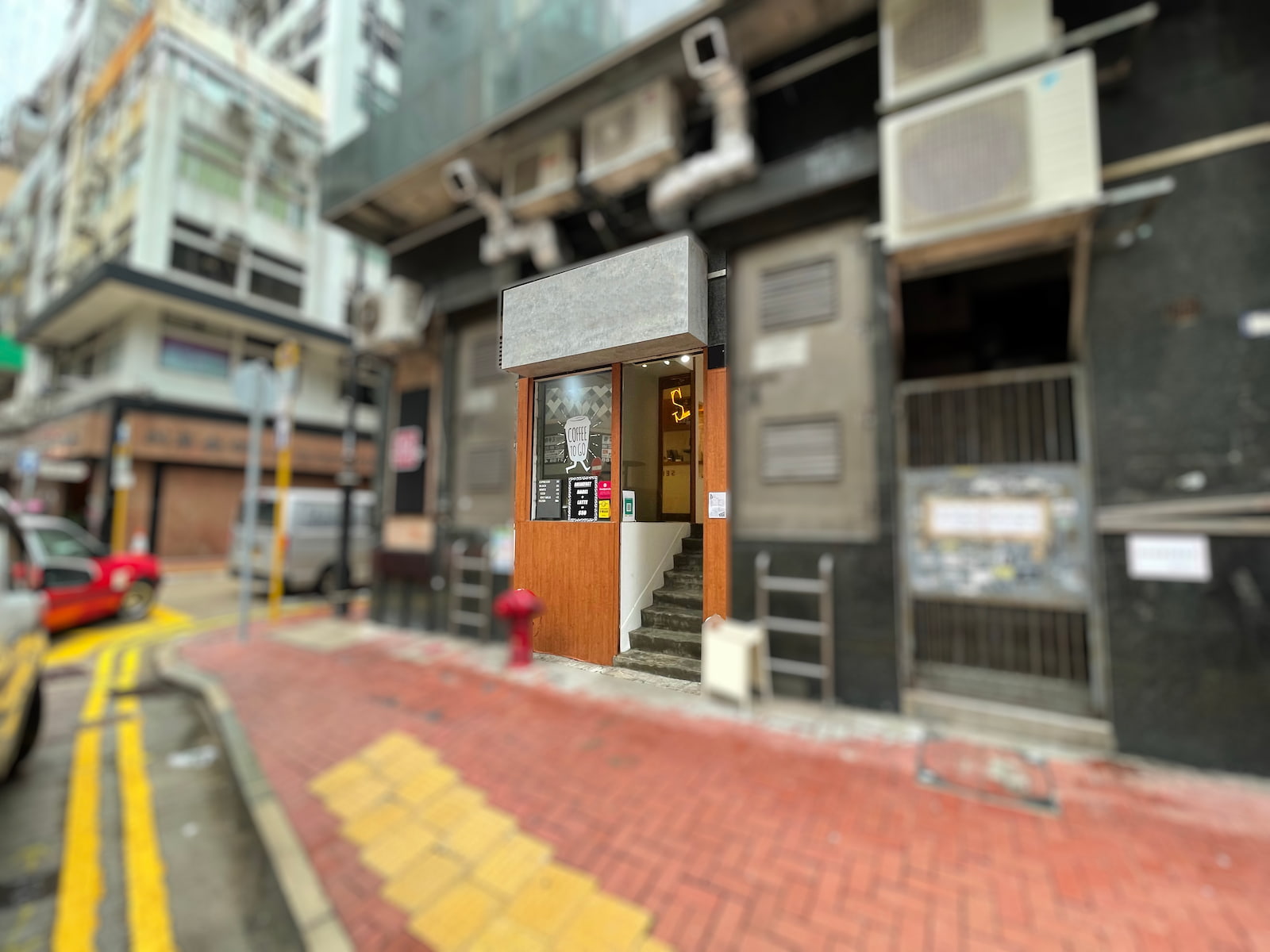 Ref#3129: A compact storefront with a wooden entrance and blank gray sign, ideal for office or shop rental, is located on a city street corner with red brick pavement, tall buildings, and visible air conditioning units.