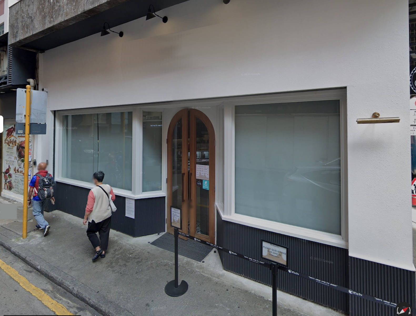 Street view of Ref#3258a modern storefront with large windows, a curved wooden door, and minimal signage. Ideal for shop or restaurant rental. Two people walk by on the sidewalk; a street pole stands to the left.