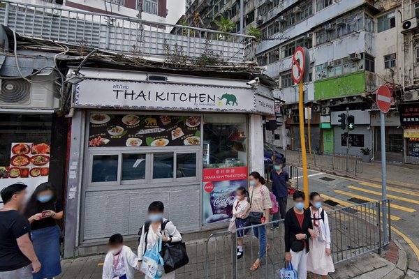 A group stands outside Ref#3394, a small restaurant for rent on a busy street corner, with menu photos displayed out front and tall buildings behind.