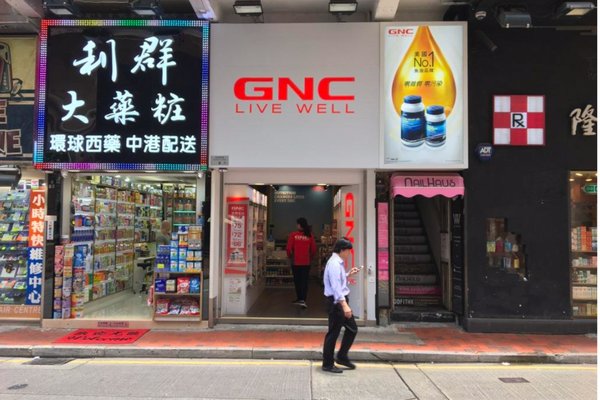A person walks past Ref#3139, a GNC store with a bright red "GNC LIVE WELL" sign, located between a pharmacy with Chinese signage and the narrow entrance of a beauty shopideal for restaurant or retail rental.