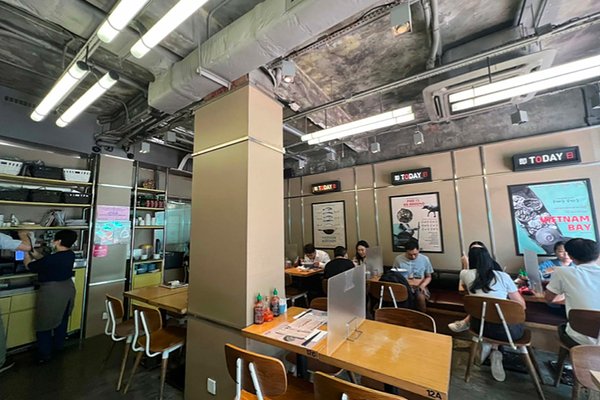 The modern interior of Ref#3221 features wooden tables, chairs, digital menu boards, and posters. Overhead lighting brightens the space, where diners are seated and a staff member works near the kitchen1perfect for restaurant rental.