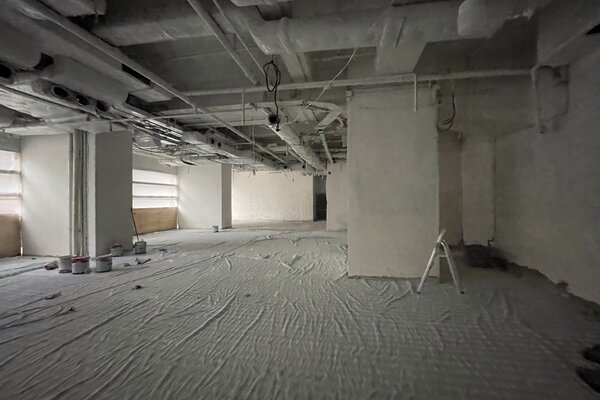 Ref#3495: Spacious, unfinished room for rent2094ideal for a restaurant, shop, or office2094with covered floors, exposed ceiling pipes, paint cans, and a white ladder near a pillar. Natural light enters through frosted left-side windows.