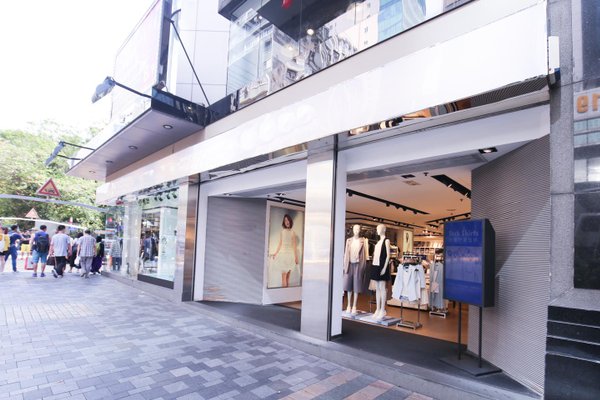 佐敦彌敦道舖位。服裝店設開啟的玻璃門及人體模型,外面佐敦彌敦道有種植樹木的街道,行人步行其中。