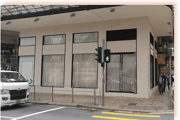 Ref#3564: Beige vacant storefront with large paper-covered windows, perfect for shop or restaurant rental. Located on a busy street corner with a lit green crossing signal, white van parked nearby, and pedestrians passing by.