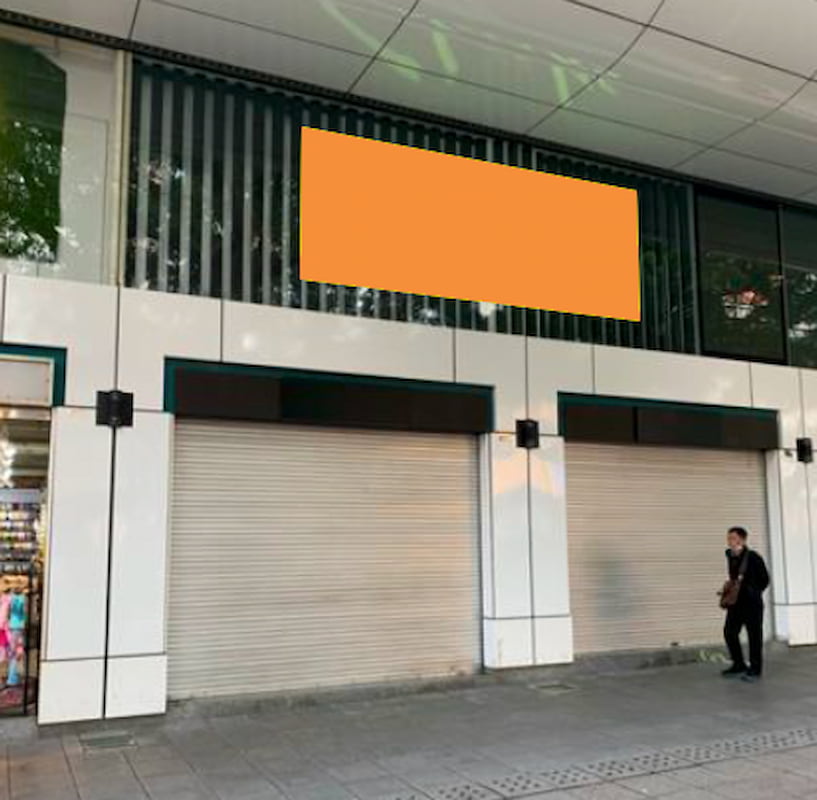 Ref#3190: Storefront with two closed metal roller shutters, large orange blank sign aboveideal for shop or restaurant rental. Person walking on the sidewalk in front of the building.