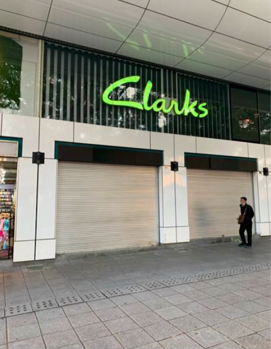 Ref#3190: Closed Clarks store with beige shutters and a bright green sign above the door; ideal rental opportunity. A person in dark clothing stands to the right on the sidewalk.