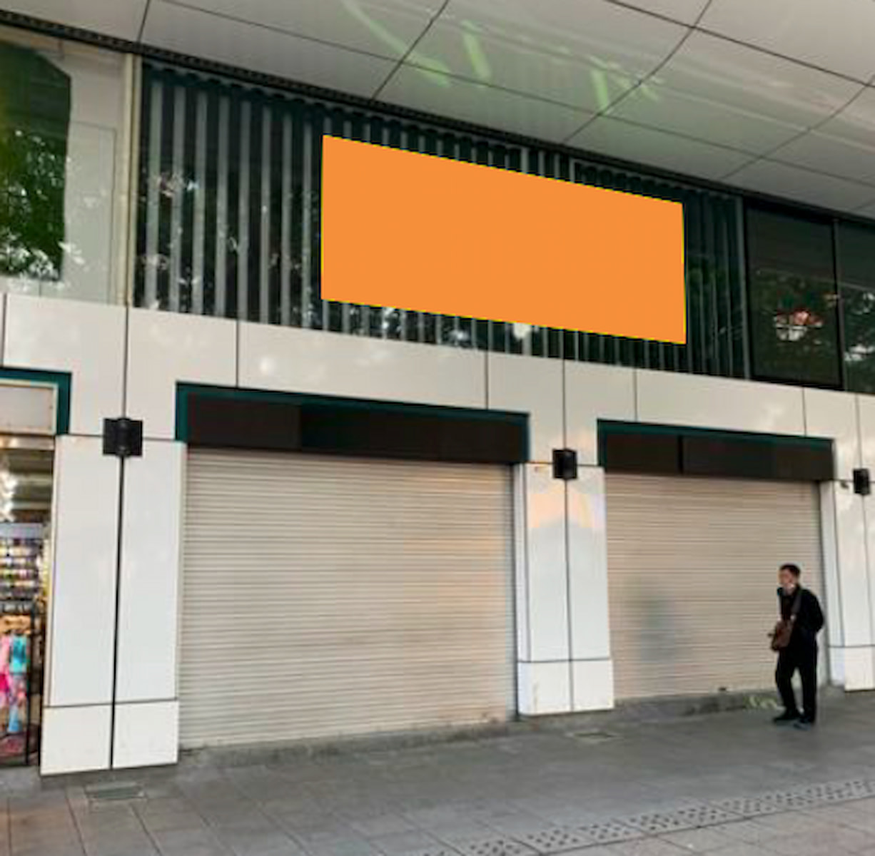 A man stands on the sidewalk in front of Ref#3190, a building with two closed roller shutters and a large blank orange sign above, indicating it's available for rent as a shop or restaurant.