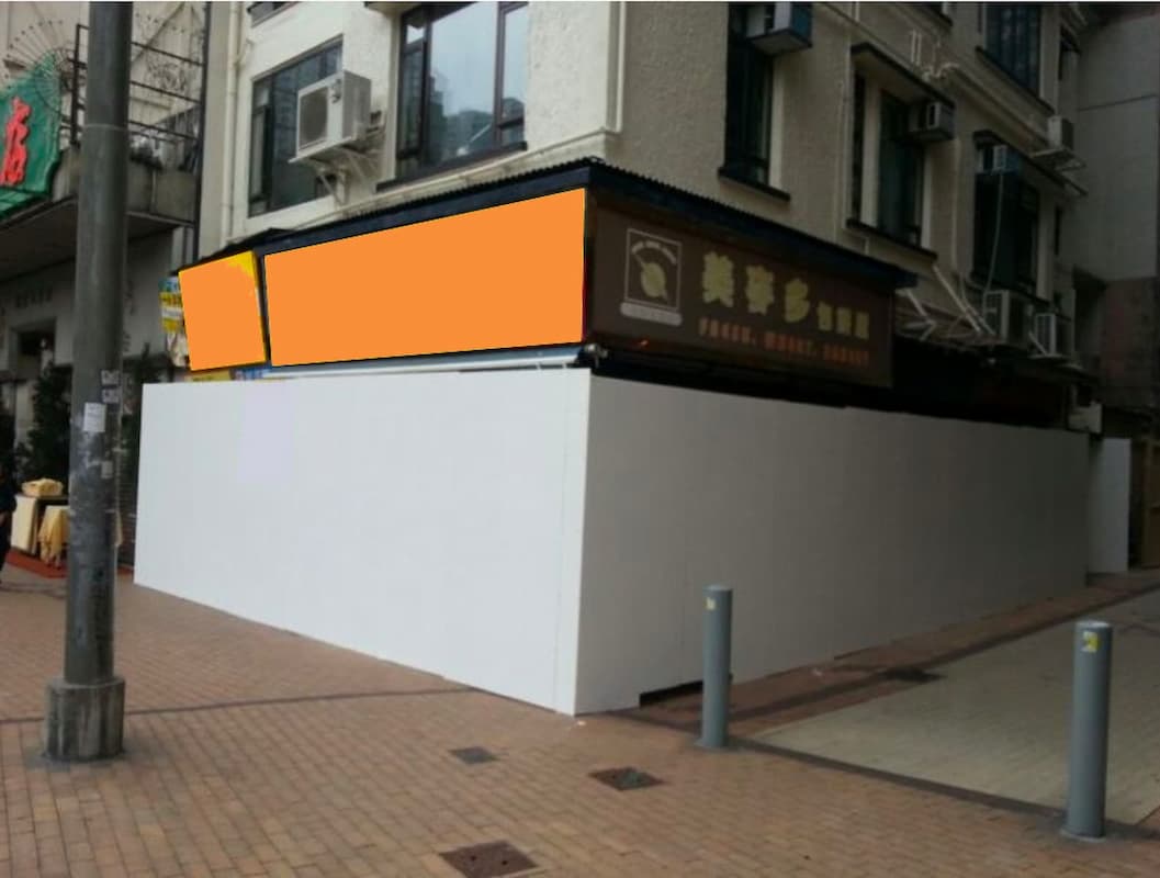Ref#3565: A corner building with a restaurant rental inside, partly obscured by tall white construction barriers. The restaurant	9s orange-blurred sign is partially visible. The sidewalk outside remains empty.