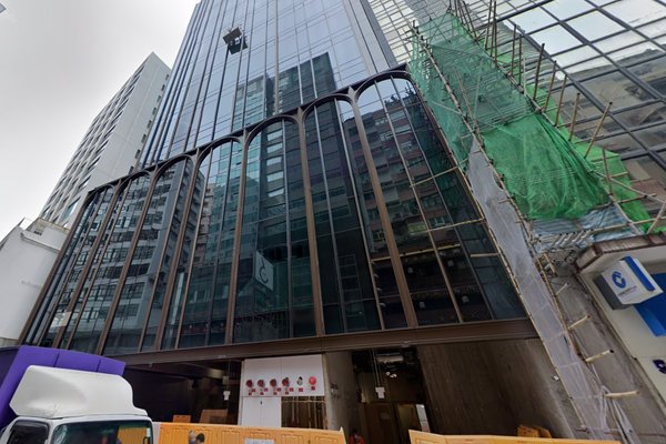 Ref#3525: A modern glass office building under construction, with scaffolding and green netting on the right and construction barriers near the entrance; surrounded by tall buildingsideal for future office or shop rental.