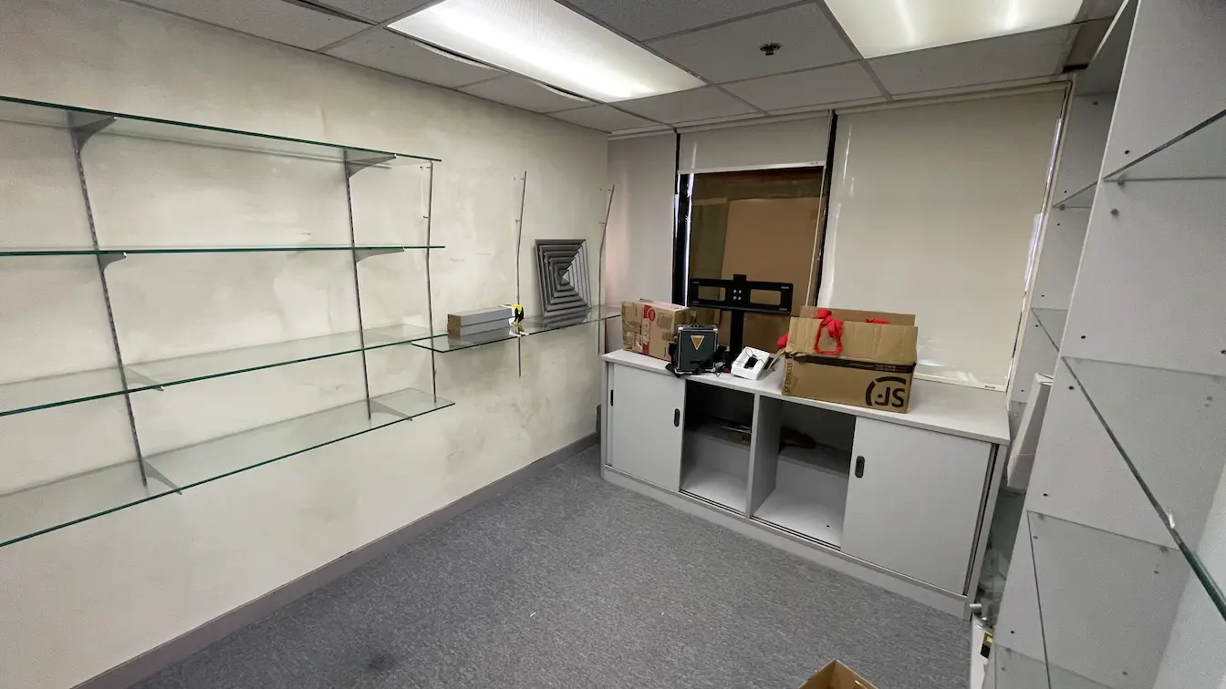Ref#3302: Small, unfurnished office for rent featuring glass wall shelves, a cabinet storing phones and boxes under the window, overhead fluorescent lights, carpeted floor, and plain walls—ideal as an office or shop space.
