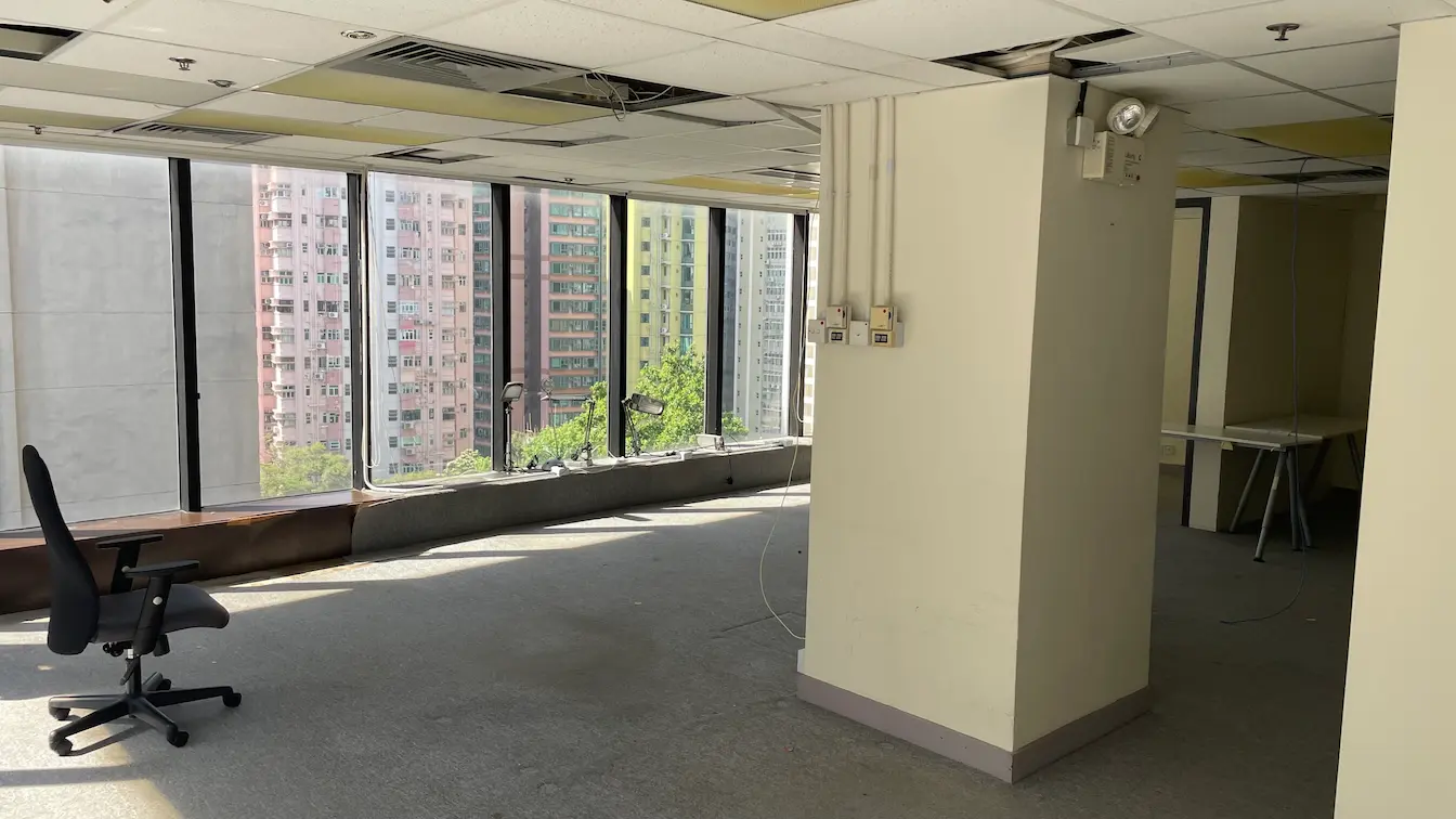 Ref#3302 offers a mostly empty office space with large windows, an office chair, a column with electrical panels, and tables in the back. Enjoy views of tall apartment buildings—ideal for those seeking spacious offices to rent.
