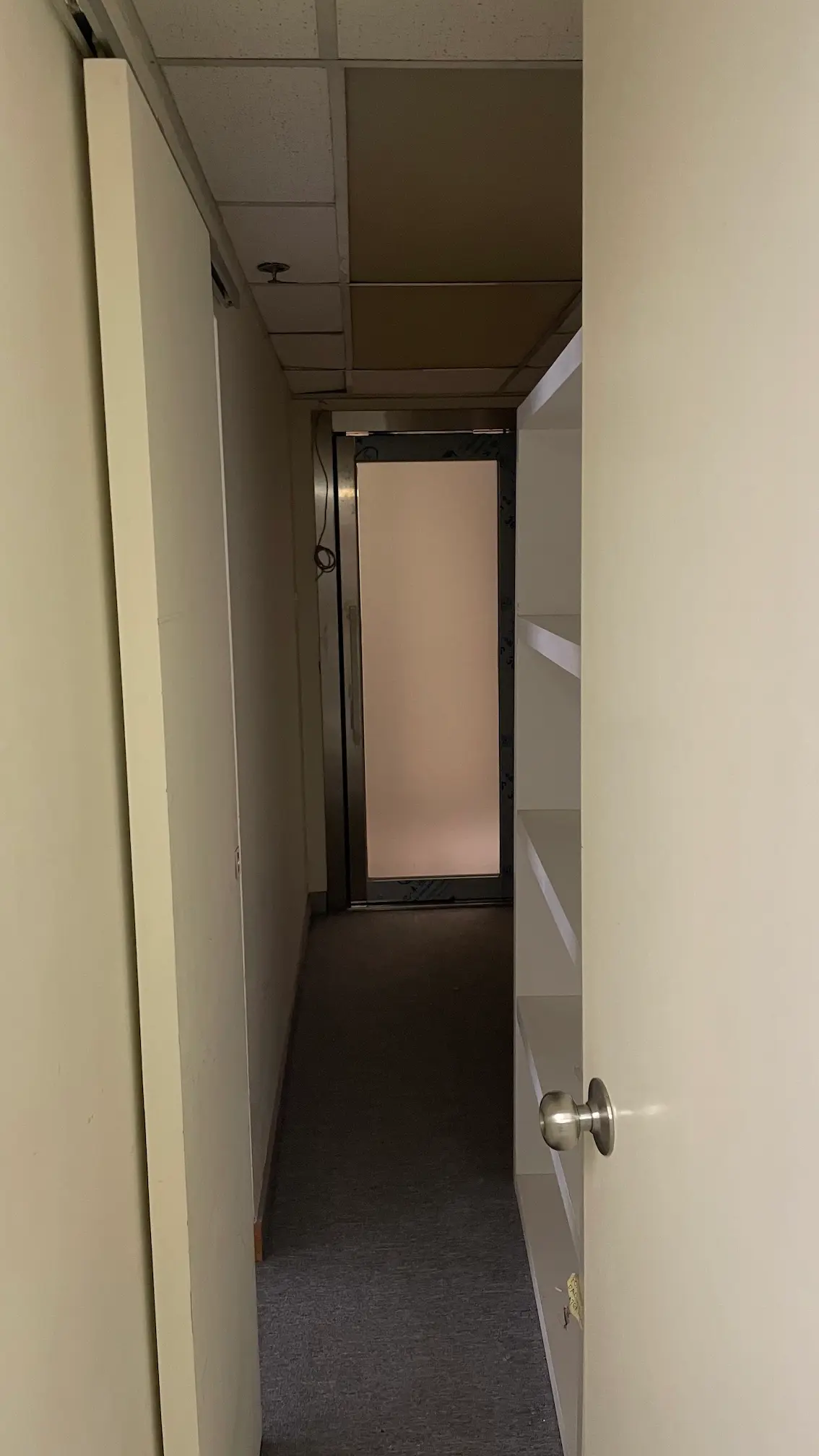 A partially open door reveals a narrow hallway with beige walls, carpeted floor, and built-in shelves—ideal for office rental (Ref#3302). At the end, there’s a closed metal door with a frosted panel.
