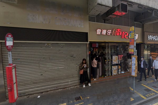 Ref#3223: A closed currency exchange shop with a metal shutter, adjacent to a restaurant with bright signage, stands near a bus stop where several people are gathered on the city sidewalk. Suitable for rent as a shop or office.