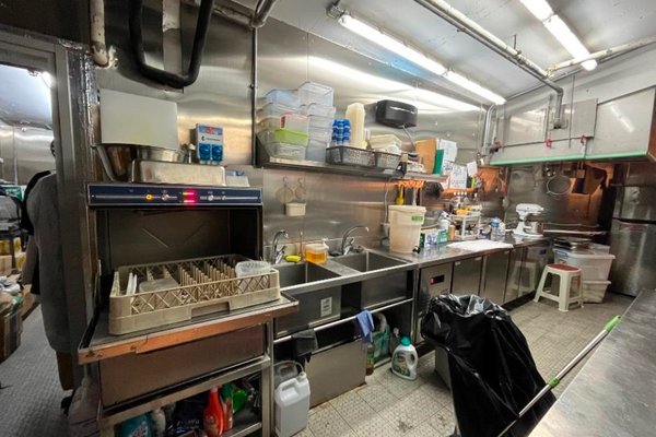 Ref#3312 is a commercial kitchen for rent, equipped with a dishwasher, sinks, shelving with containers, cleaning supplies, utensils, stainless steel surfaces, and white tiled floors in an organized and well-lit space.