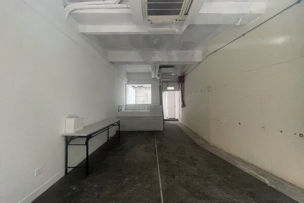 Ref#3584: A long, narrow room with white walls, exposed pipes, a side table, and back counter by a doorway. The worn floor makes it ideal for shop or restaurant rental.