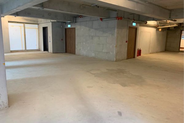Ref#3380: Spacious, empty concrete room with unfinished walls and ceilingideal to rent as an office, shop, or restaurant. Features several closed wooden doors, emergency exit signs, and fire extinguishers in a brightly lit space.