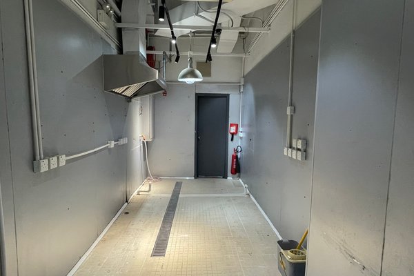 Ref#3579: Clean, empty industrial room with gray walls, exposed pipes, floor drain
ideal for shop or office rental. Includes wall-mounted outlets, metal vent hood, overhead lights, fire extinguisher, and mop bucket in the corner.