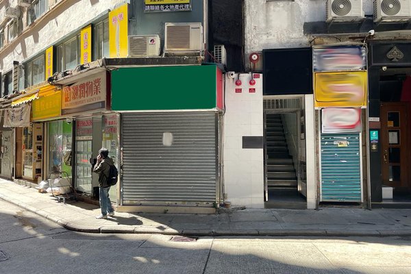 A person stands on a sunny sidewalk beside closed shops with metal shutters and faded signs, some available to rent as Ref#3501 for a shop or restaurant. A staircase is between two storefronts, with air conditioners mounted above.