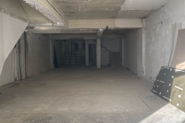 Ref#3591: Spacious unfinished basement with concrete floors, exposed beams, and minimal lighting1perfect for a restaurant or creative shop rental. Ladders, boards, and construction materials are stacked along the back and right wall.