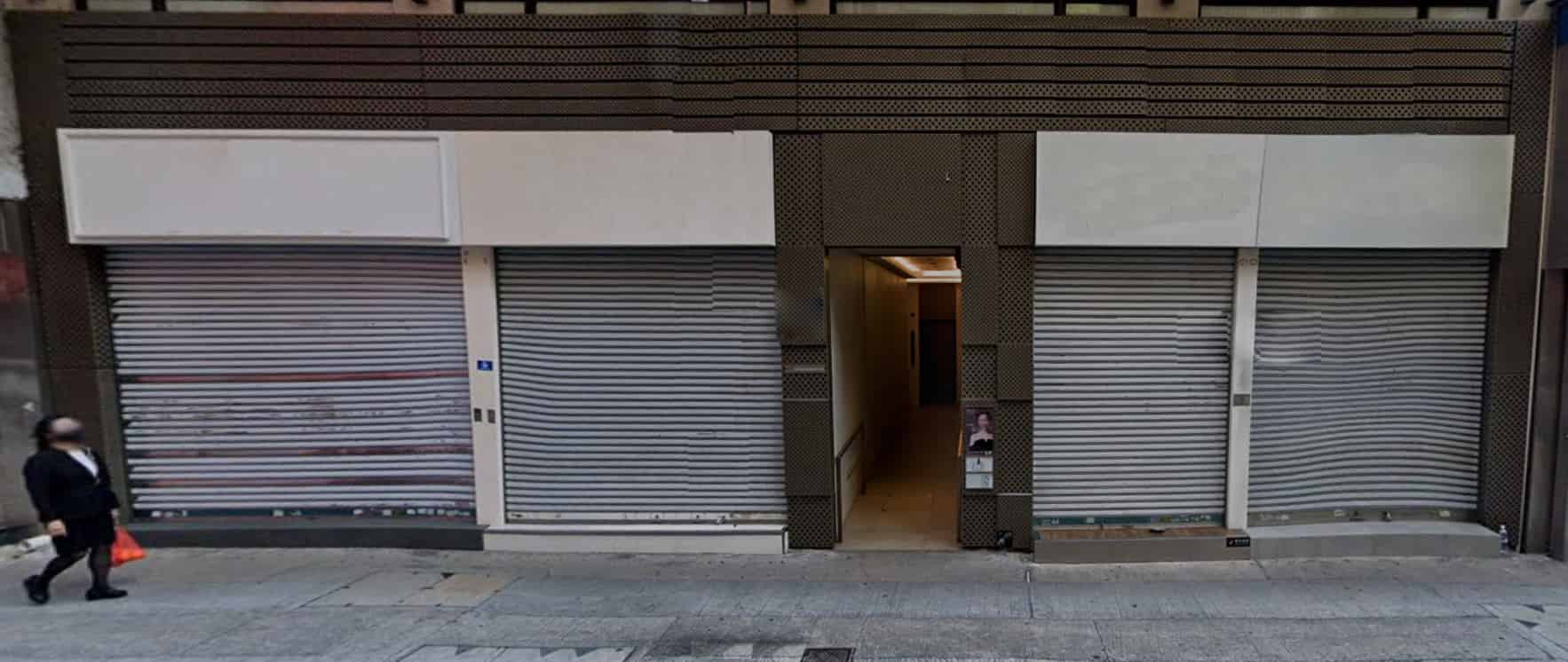 A person walks past two closed storefronts with metal shutters and a narrow lit entrance between them on a sloped city street—Ref#3247, ideal for shop or office rental.