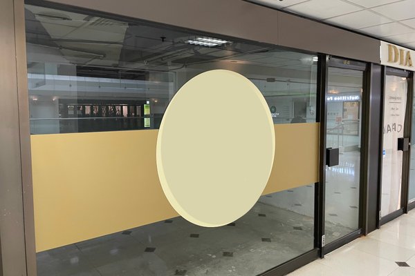 Ref#3170: A closed glass storefront with a beige horizontal stripe and large circle at the center. Ideal for renting as shop or office space, it features an empty interior reflecting ceiling lights and the mall corridor.