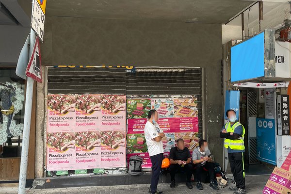 Five people, some in reflective vests, gather on a sidewalk next to a closed rent shop named Ref#3207. The shutter is covered with Foodpanda and PizzaExpress ads, and some are holding drinks or food.