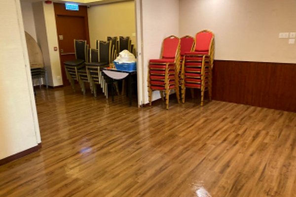 Ref#3156: A clean, unused space ideal for a shop or office, with wooden flooring, stacks of red cushioned chairs, gray chairs in the corner, and a small table with a white cloth near the doorway.
