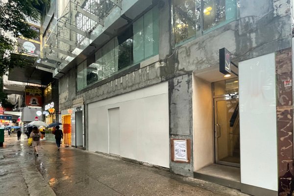 On a rainy city day, people walk under umbrellas past Ref#3198the building on the right with unfinished walls, large glass windows above, and a lit entranceideal for restaurant rental. The wet street and sidewalk reflect the rain.
