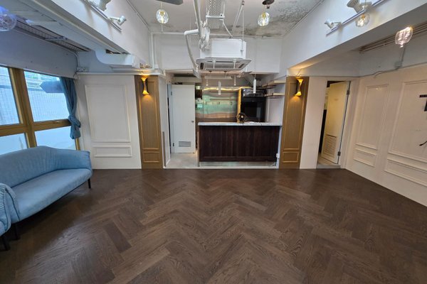 Ref#3381: Modern open-concept space for office or shop with dark herringbone wood floors, a small kitchen with dark cabinets and stainless steel appliances, white walls, and a light blue sofa near large windows with blue curtains.