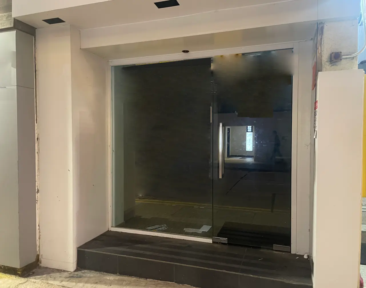 Ref#3459 features a clear glass door with metal handles at the entrance, ideal for shop or office rentals. Surrounded by white walls and black floor tiles, its empty interior provides a blank slate, with shimmering reflections on the glass.